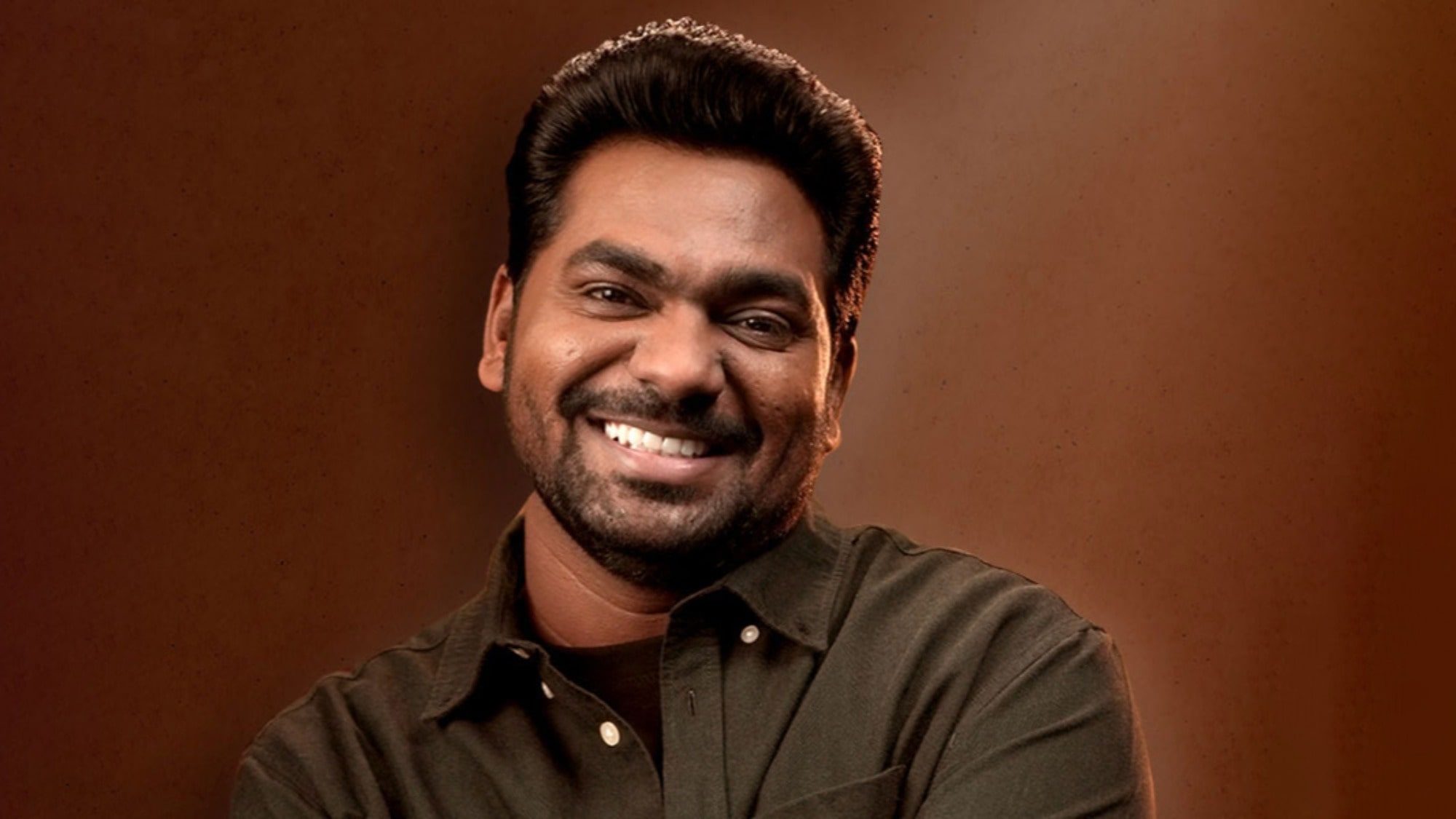 Zakir Khan smiling at the camera with a brown background.