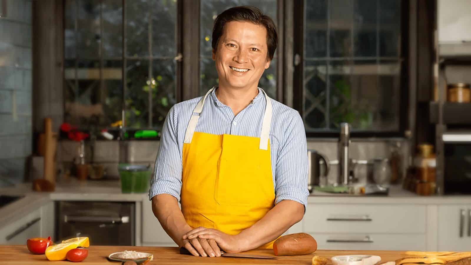 16x9 A smiling person wearing a blue-striped shirt and a yellow apron stands at a kitchen counter, engaged in Kitchen Conversations with various food items, inspired by J. Kenji López-Alt.