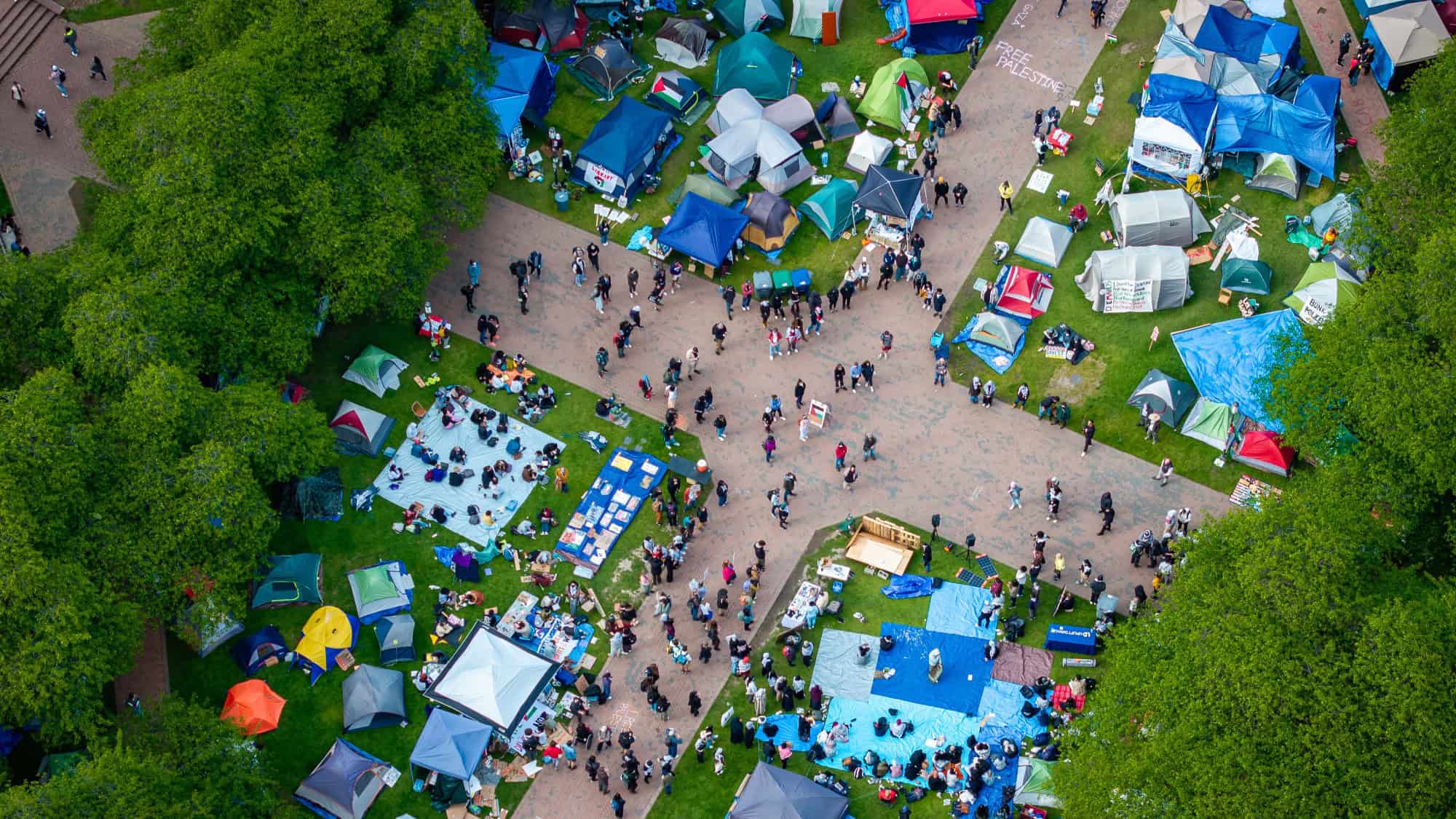 240507_SeanFan_PopularUniversity_Drone_CMS-11-16-9-2000x1125 An aerial view of a park with people gathering around various tents and blue tarps in different sections, bordered by green trees. The scene suggests an outdoor event or festival, reminiscent of an encampment deal fostering a range of opinions and ideas.