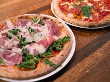 Bar Cotto-4-3-360x270-min Two pizzas on wooden tables; one topped with prosciutto, arugula, and shaved parmesan, the other with tomato, mozzarella, and basil at a culinary retreat.