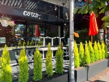 Cotto Belltown-4-3-360x270-min Outdoor dining area of a restaurant named "cotto" with vibrant green shrubs, white chairs, and red umbrellas under a black awning at a culinary retreat.