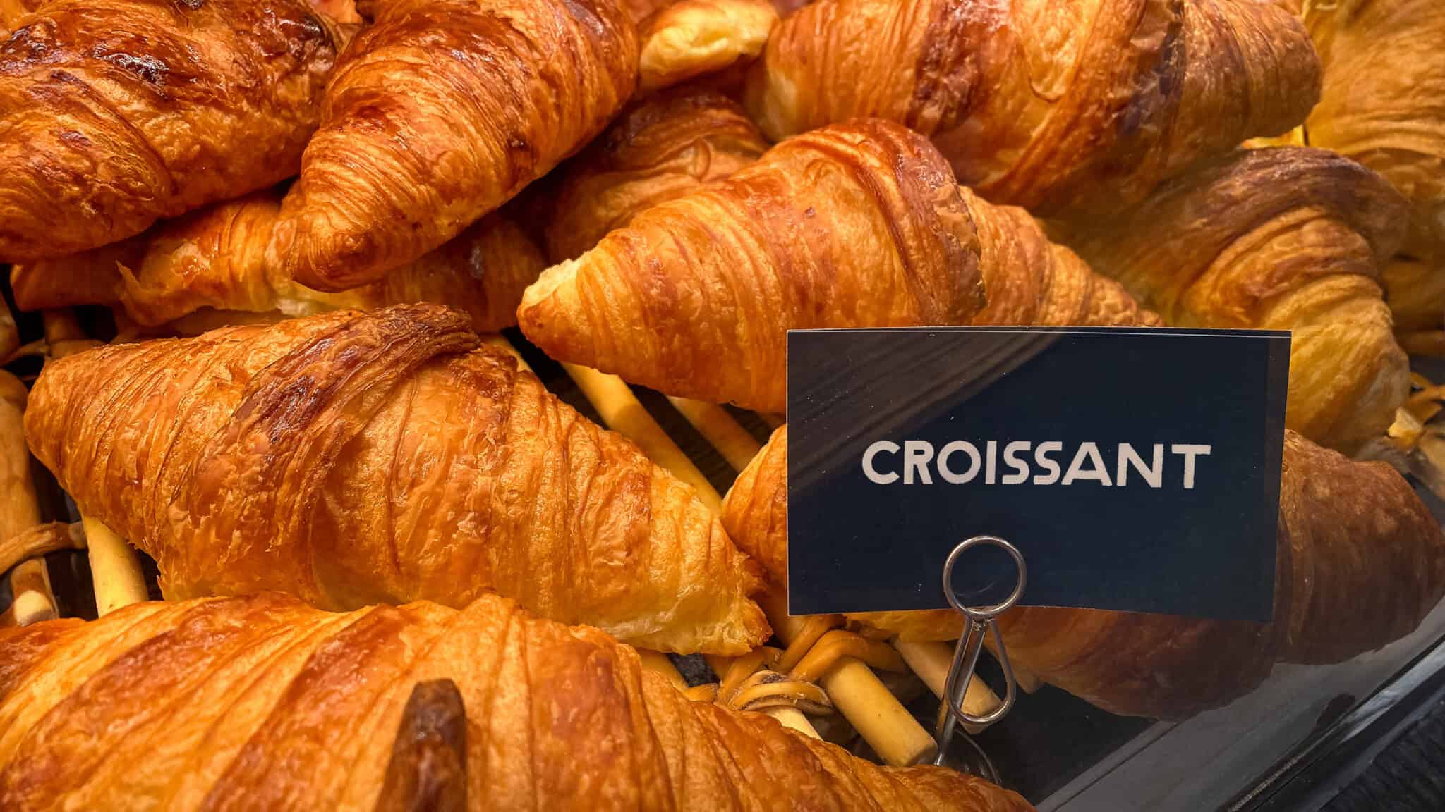 Croissant_16x9 A display of golden-brown croissants with a black sign labeled "croissant" (pronunciation: kruh-sahnt) in a bakery case.