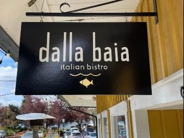 Dalla Baia-4-3-360x270-min Sign for "dalla baia italian bistro" with a gold bird icon, hanging outside a building with yellow trim, featured in Seattle magazine.