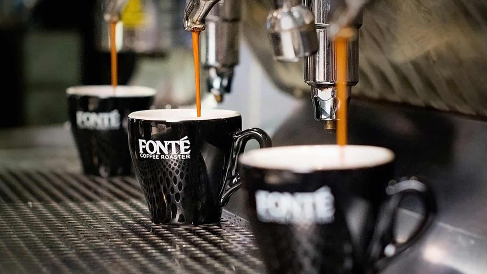 FonteEspresso_16x9 Three black Fonte coffee mugs are placed under espresso machines with espresso being dispensed into them, creating a scene of artisanal coffee art.