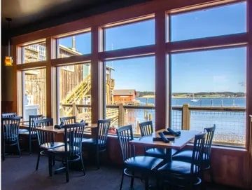 Front Street Grill Interior of a restaurant with large windows overlooking a sunny waterfront view in Seattle, featuring empty tables and chairs.