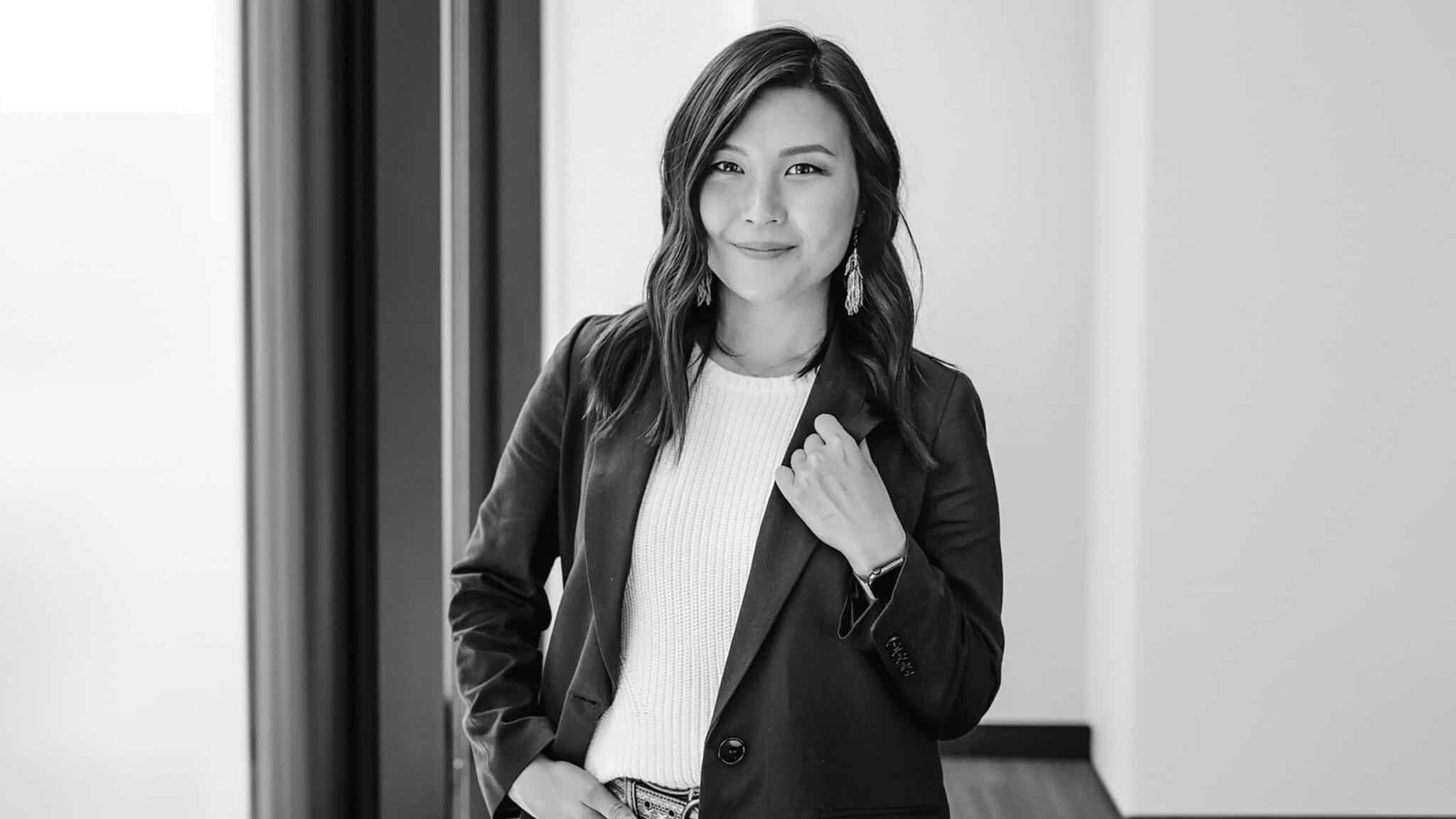 GarceYoo_16x9-min A professional black and white portrait of an asian woman smiling, standing in an office environment, dressed in a blazer and white top.
