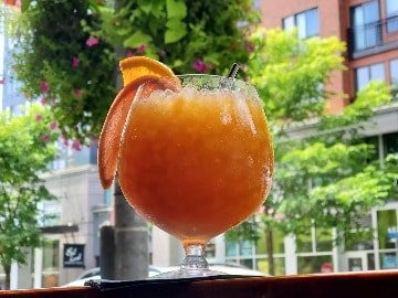 Herb and Bitter-4-3-360x270-min A chilled orange cocktail garnished with a slice of orange, served in a glass on an outdoor table with Seattle magazine in the background.