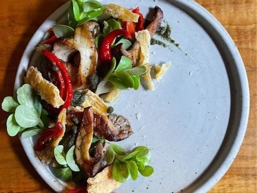 Matia Kitchen A plate with a mixed salad of leafy greens, sliced mushrooms, roasted red peppers, and crispy bread pieces, garnished with a drizzle of sauce, perfect for enjoying at one of the cozy Sheringham restaurants, on a wooden table.