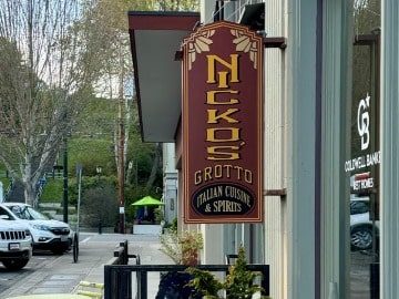 Nicko's Grotto-4-3-360x270-min A vertical street sign for "Nick's Grotto Italian Cuisine & Culinary Retreat" mounted on a post, with a street and greenery in the background.