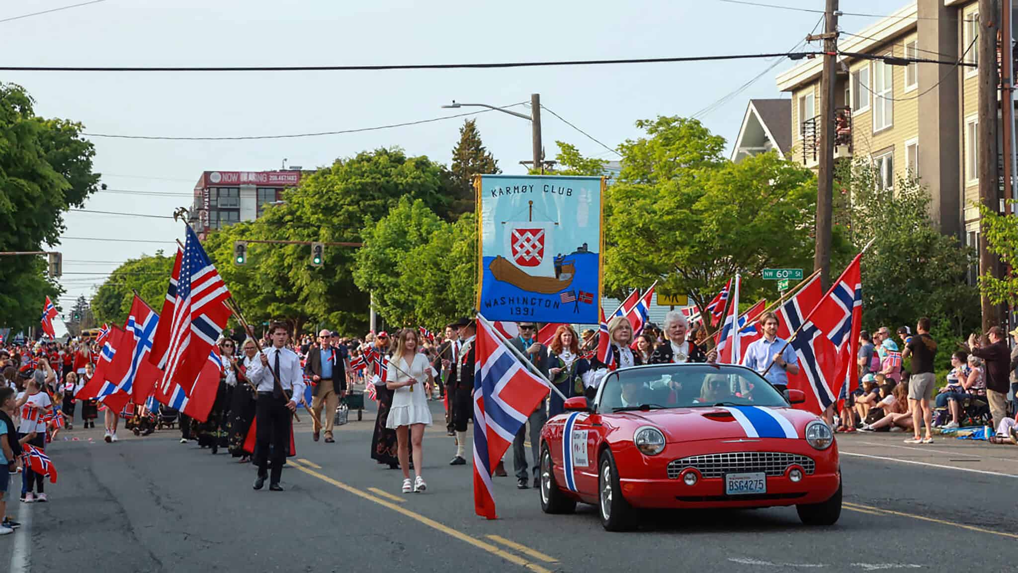 NorwegianConstituionDay_ParadeKarmoyClub_Pic03_16x9 A parade celebrating Norwegian Constitution Day features people marching with Norwegian flags. A red convertible decorated with flags leads, followed by a banner for the Leif Erikson Lodge 2-001 and other participants on a street lined with spectators. Hip hurra echoes through the crowd, making it a festive Friday event.