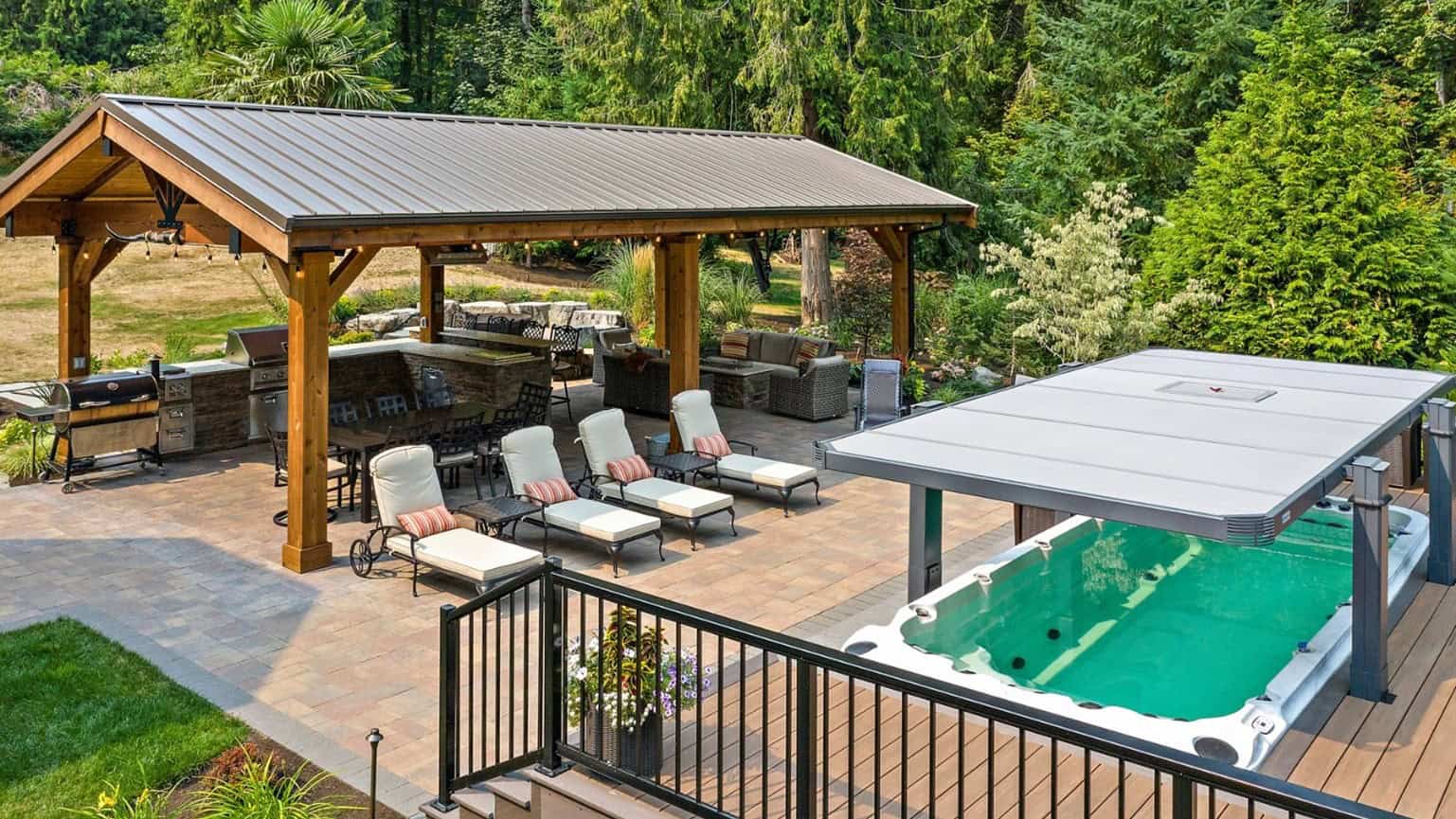 PCF_ToftProject 16x9 A backyard with a covered patio, sponsored by PCF, featuring a dining area and lounge chairs next to a swimming pool with a retractable cover, surrounded by trees and greenery.