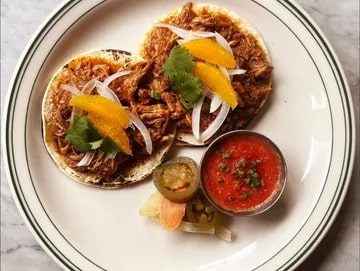 Percy's and Co Three chicken tacos topped with onion and cilantro, featured in Seattle magazine, served with a side of salsa, pickled vegetables, and a slice of lime on a white plate.