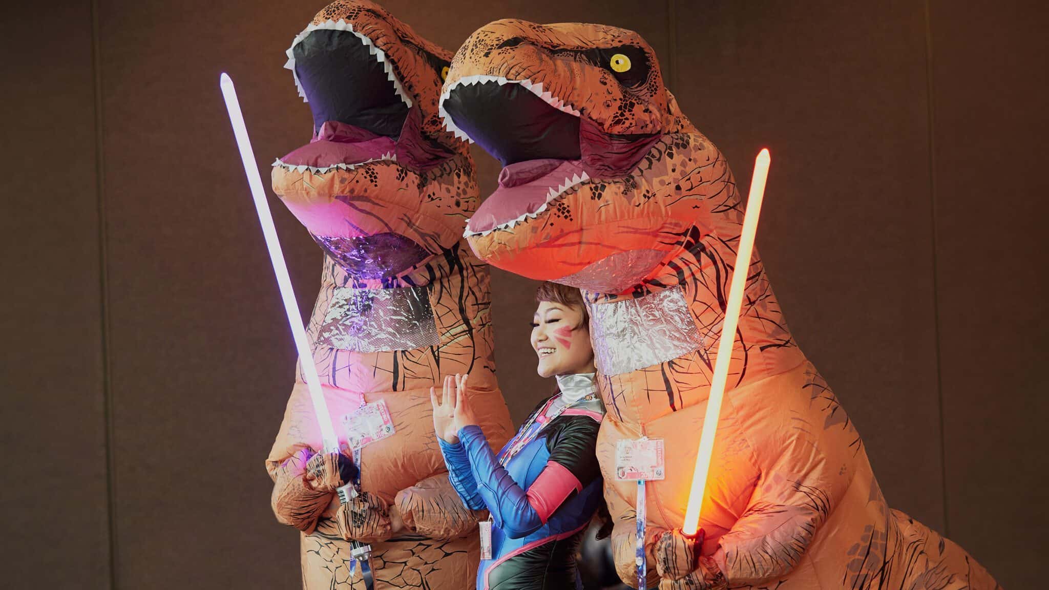 SakuraCon_16x9 Two individuals in inflatable t-rex costumes wielding lightsabers alongside a smiling person in cosplay return to Sakura-Con, Seattle.