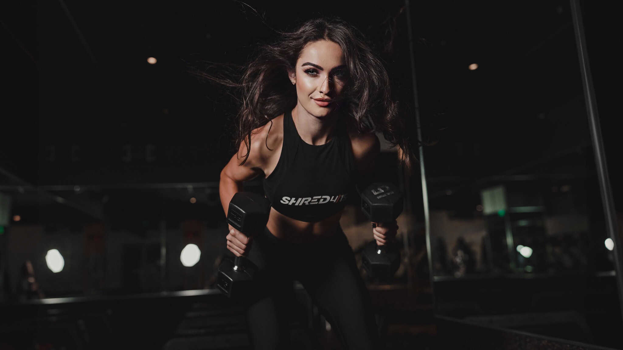 StrengthTraining_16x9 Woman exercising indoors, performing a bent-over dumbbell row, wearing exercise attire, and looking ahead with determination.