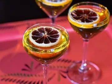 The kro bar Three cocktail glasses with a slice of dried blood orange on top, set on a pink surface with a patterned design, illuminated by warm lighting at a culinary retreat.