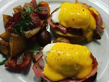 The Well and Table A plate of eggs benedict with Canadian bacon and hollandaise sauce, featured in Seattle magazine, accompanied by a side of seasoned roasted potatoes.