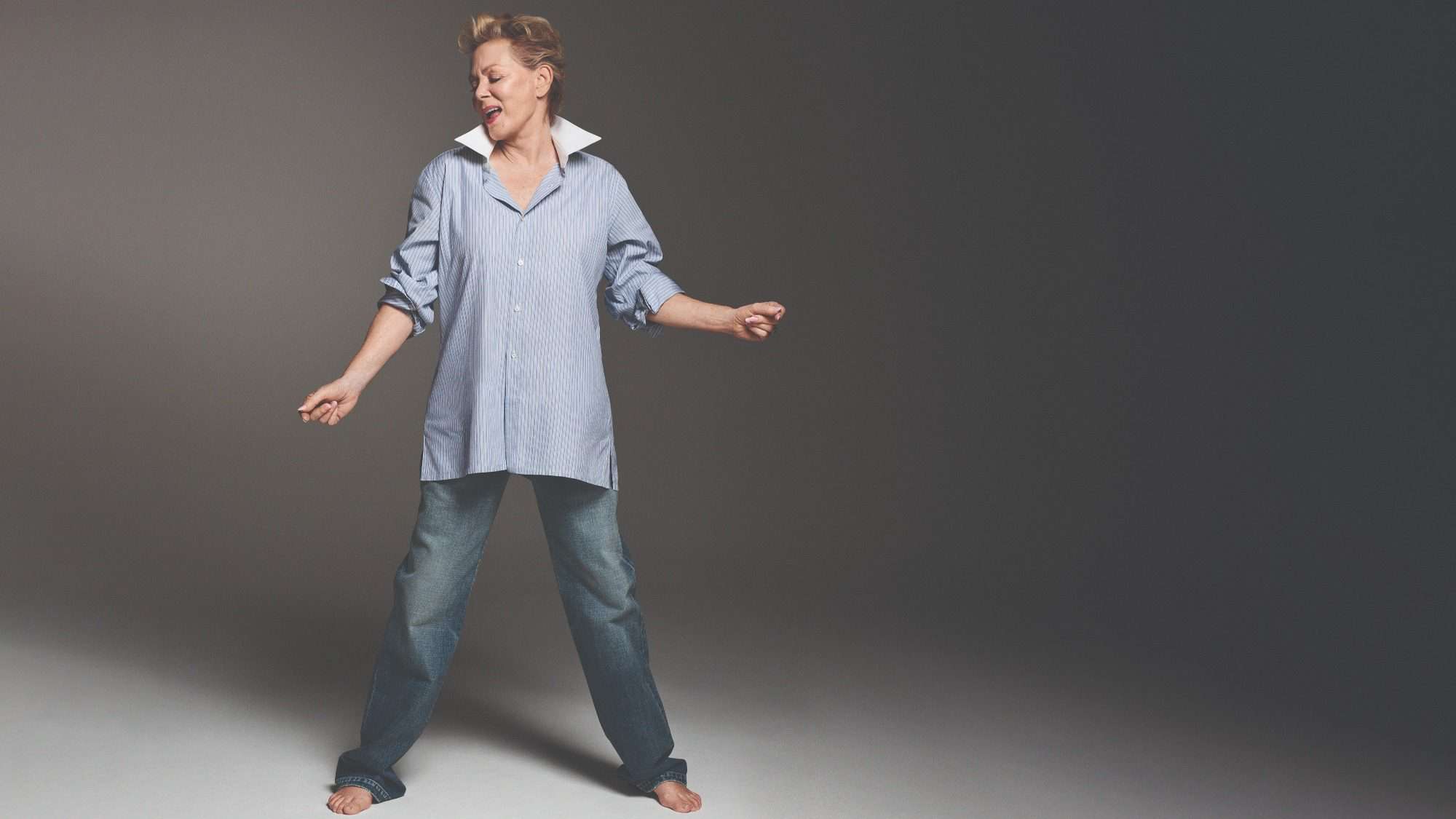 tw_jean_smart-16-9-2000x1125-min A middle-aged actress dancing joyfully in a studio, wearing jeans and an oversized shirt, with a subtle smile on her face.