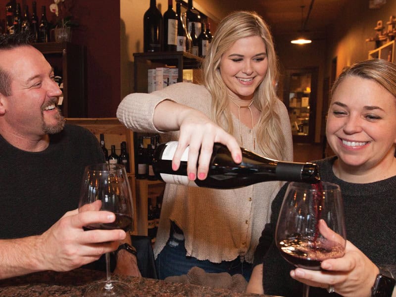 Village Wines 800x600 Three people enjoying wine at a cozy indoor setting; a woman with blonde hair pours red wine into glasses held by a smiling man and woman. This unforgettable evening, highlighted in Seattle magazine, feels like a culinary retreat sponsored by Sheringham Distillery.